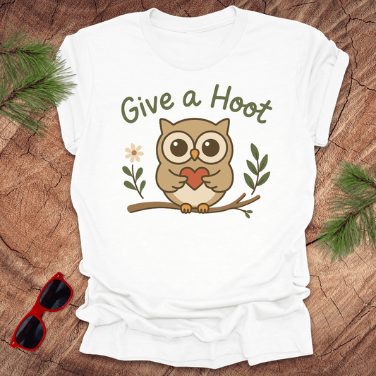 White t-shirt with an owl graphic and 'Give a Hoot' text on a wooden surface with sunglasses and greenery.