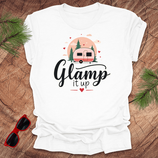 White t-shirt with 'Glamp it up' design featuring a pink camper on a wooden surface with sunglasses and greenery.