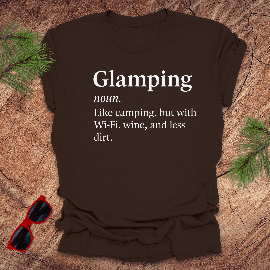 Brown t-shirt with 'Glamping' definition on a wooden surface with sunglasses and pine branches.