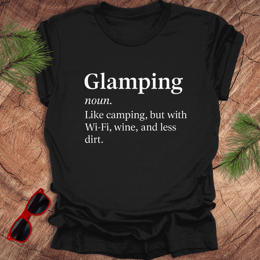 Black t-shirt with 'Glamping' definition text on a wooden surface with sunglasses and pine branches.