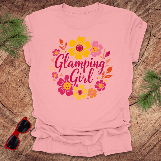 Pink t-shirt with 'Glamping Girl' text and floral design on a wooden surface with sunglasses and greenery.