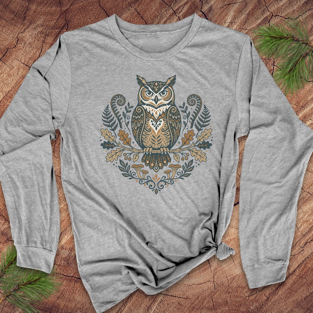 A heather gray long-sleeve t-shirt with a detailed owl graphic on the chest, styled flat on a rustic wood background with pine needles.