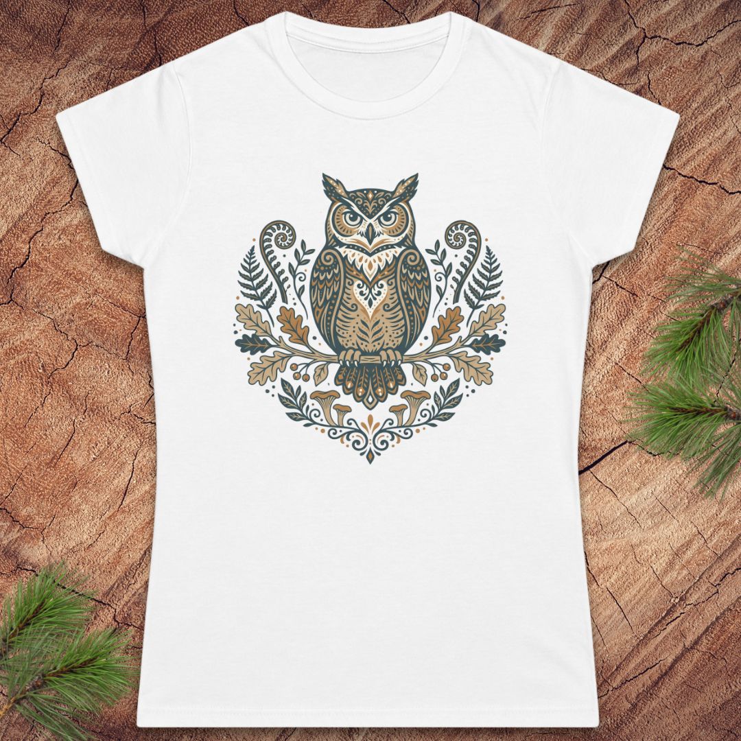 A white short-sleeve crewneck t-shirt featuring a detailed graphic of an owl surrounded by leaves and ferns in earthy brown, beige, and muted green tones. The t-shirt is displayed flat on a textured wood background with pine branches in the corners.
