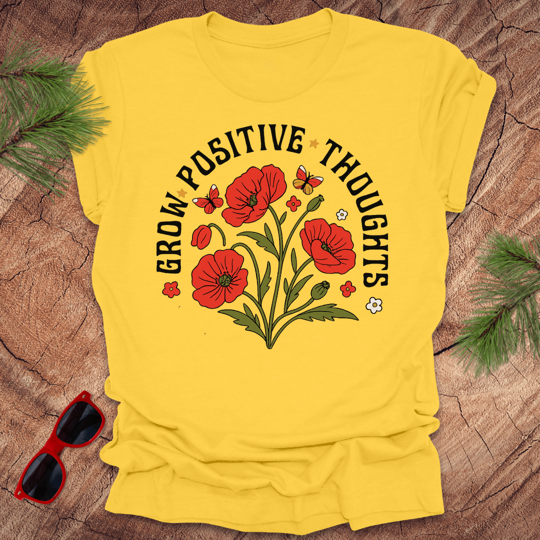 Yellow t-shirt with red poppies and 'Grow Positive Thoughts' text on a wooden surface with sunglasses.