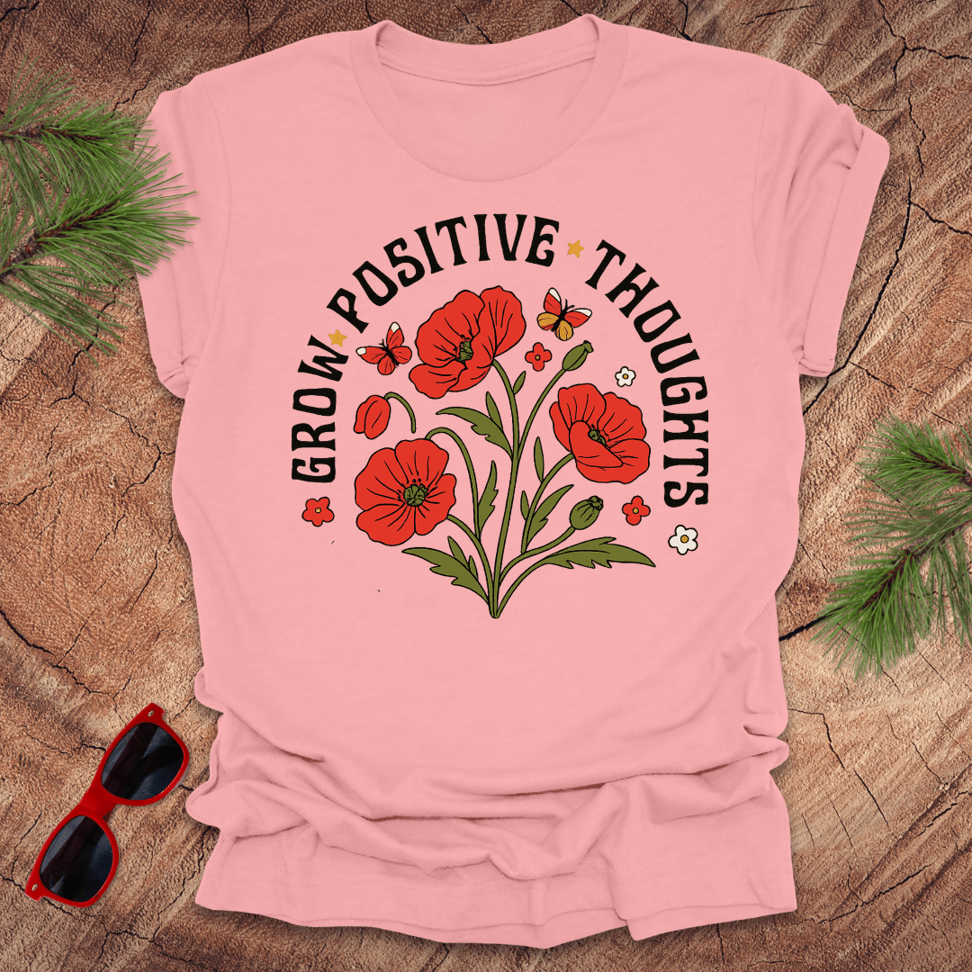 Pink t-shirt with red floral design and text on a wooden surface with sunglasses and greenery.
