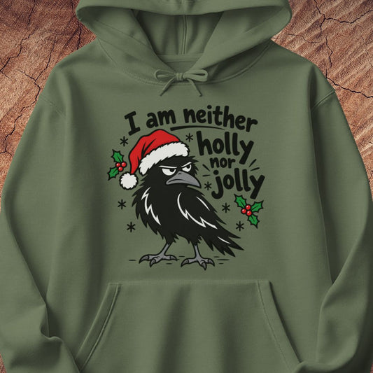 Military green hoodie with a grumpy raven in a Santa hat surrounded by holly and snowflakes and the words I am neither holly nor jolly, laid flat on a rustic wood background.