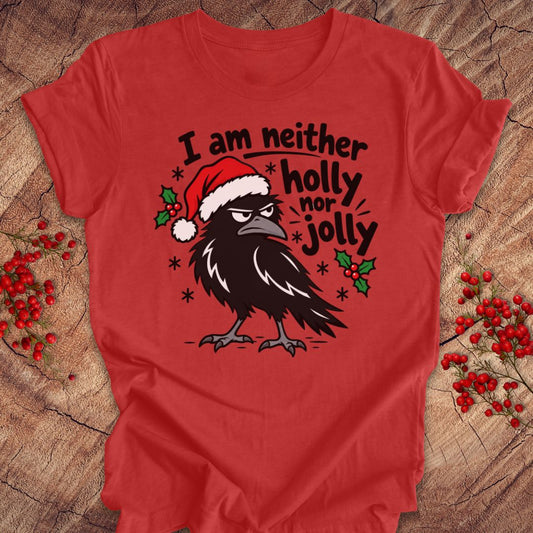 Heather red t-shirt with a grumpy raven in a Santa hat surrounded by holly and snowflakes and the words I am neither holly nor jolly, laid flat on a rustic wood background with red winter berries.