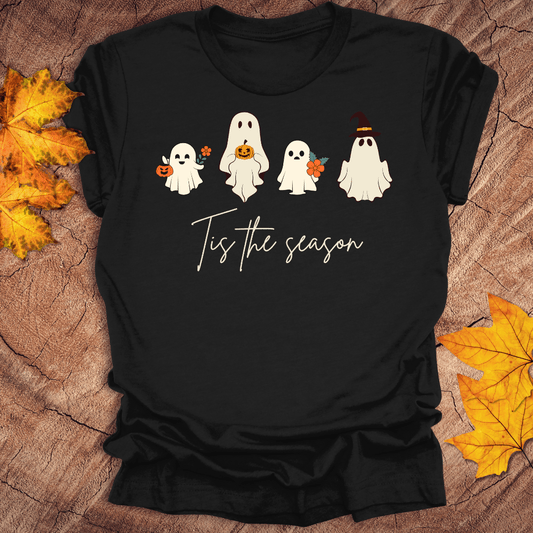 Black t-shirt with Halloween-themed design featuring ghosts and text 'Tis the season' on a wooden surface with fall leaves.