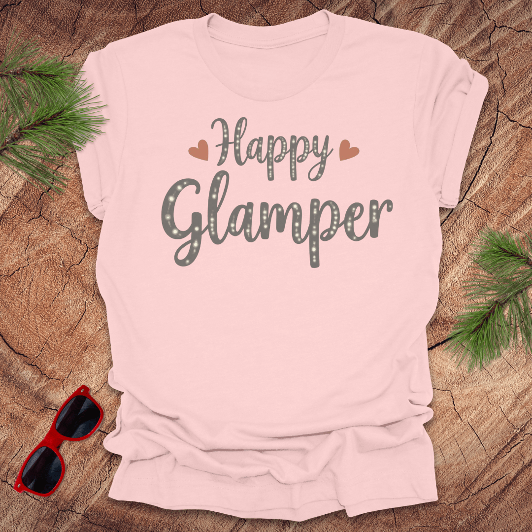 Pink t-shirt with 'Happy Glamper' text on a wooden surface with sunglasses and greenery.