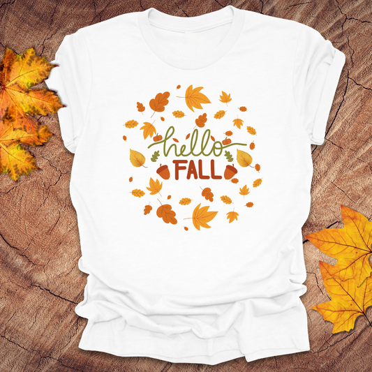 White t-shirt with 'hello FALL' text and autumn leaf design on a wooden surface with fall leaves.
