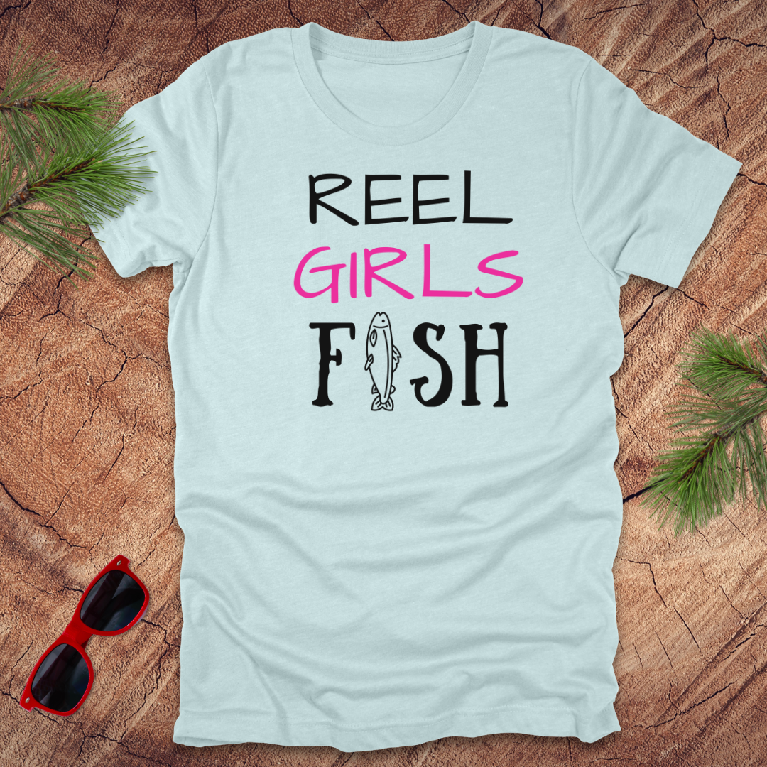 Light blue t-shirt with 'Reel Girls Fish' design on a wooden surface with sunglasses and greenery.