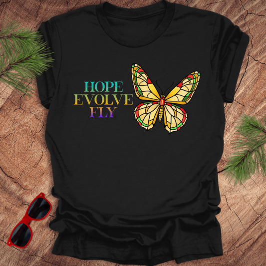 Black t-shirt with a colorful butterfly graphic and text on a wooden surface with sunglasses and pine branches.