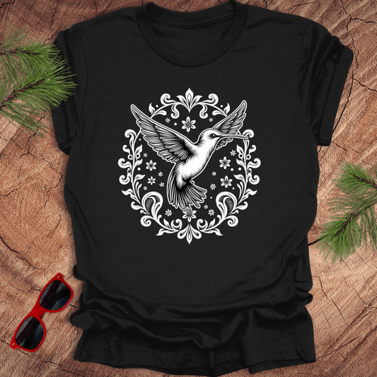 black t-shirt with a graphic of a hummingbird surrounded by floral and ornamental designs, displayed on a wooden surface with pine needles and red sunglasses.
