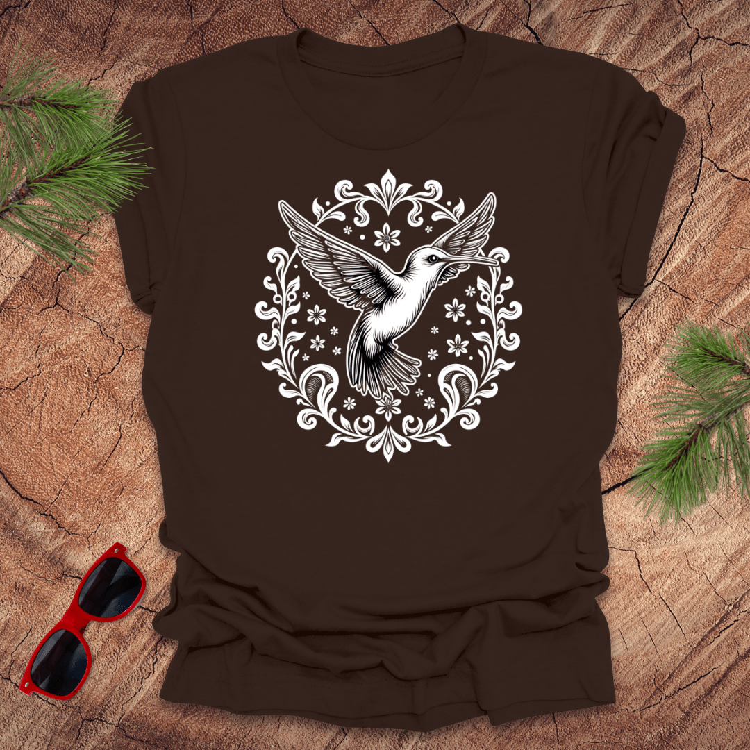 brown t-shirt with a graphic of a hummingbird surrounded by floral and ornamental designs, displayed on a wooden surface with pine needles and red sunglasses.