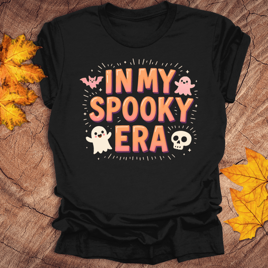 Black t-shirt with 'In My Spooky Era' text and Halloween graphics on a wooden surface with fall leaves.