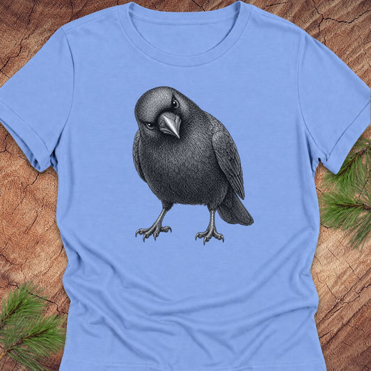 Carolina blue women's relaxed t-shirt with a pencil-style American Crow tilting its head and giving a judgy stare, laid flat on a rustic wood background with pine branches.