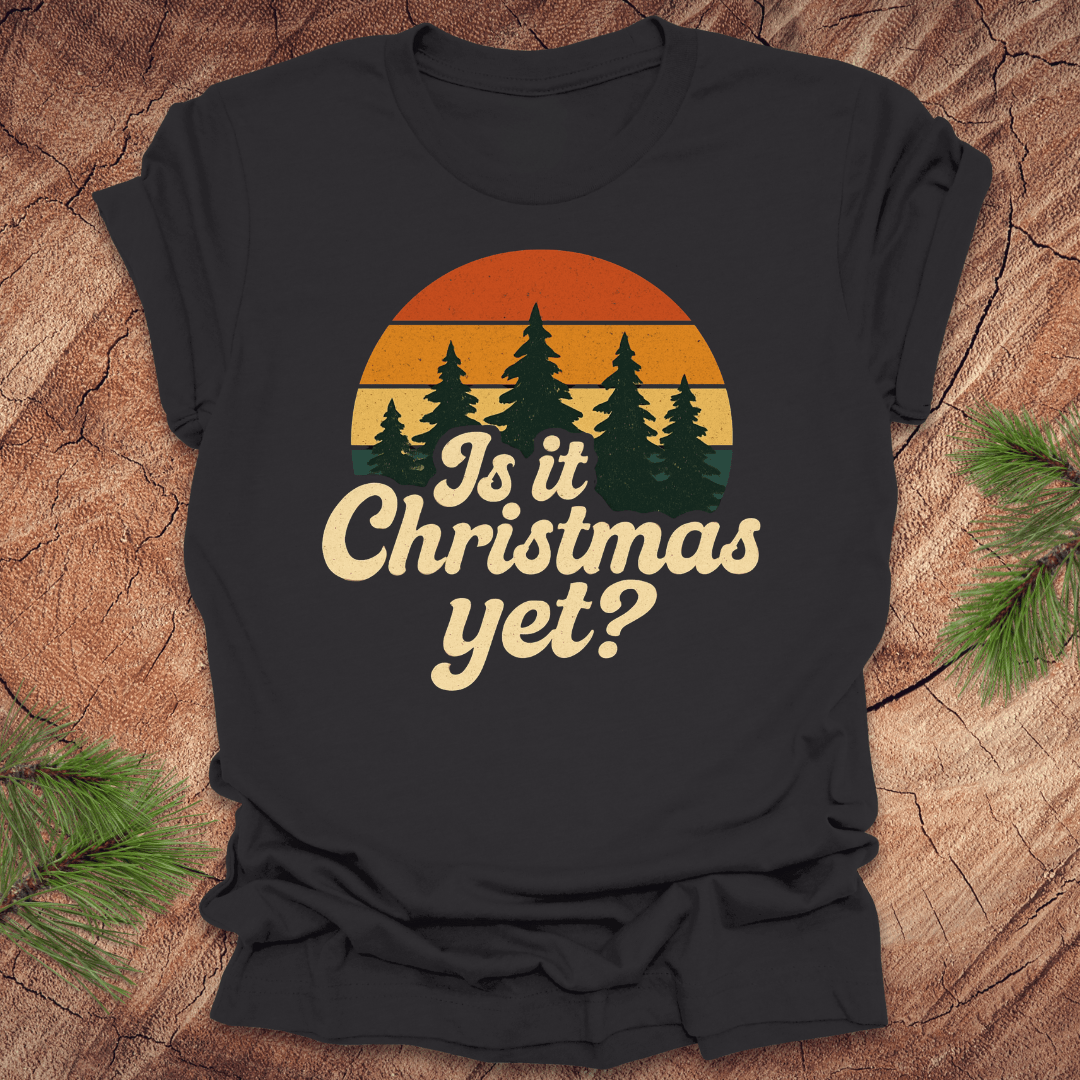 Black t-shirt with 'Is it Christmas yet?' design on a wooden surface with pine branches.