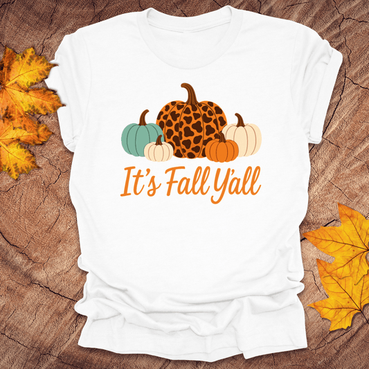 White t-shirt with pumpkins and 'It's Fall Y'all' text on a wooden surface with fall leaves.