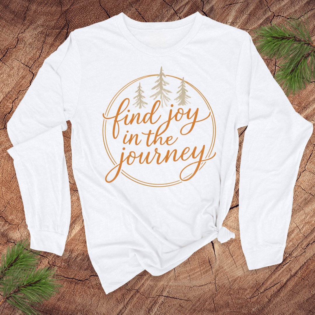 White long-sleeve shirt with 'find joy in the journey' text on a wooden surface with pine branches.