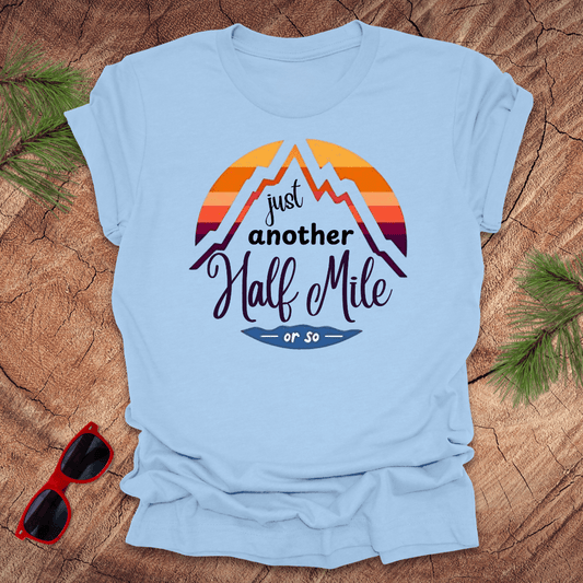 A baby blue t-shirt with a mountain graphic and the text 'Just Another Half Mile' on the front, displayed on a wooden surface with pine needles and a pair of sunglasses in the background.