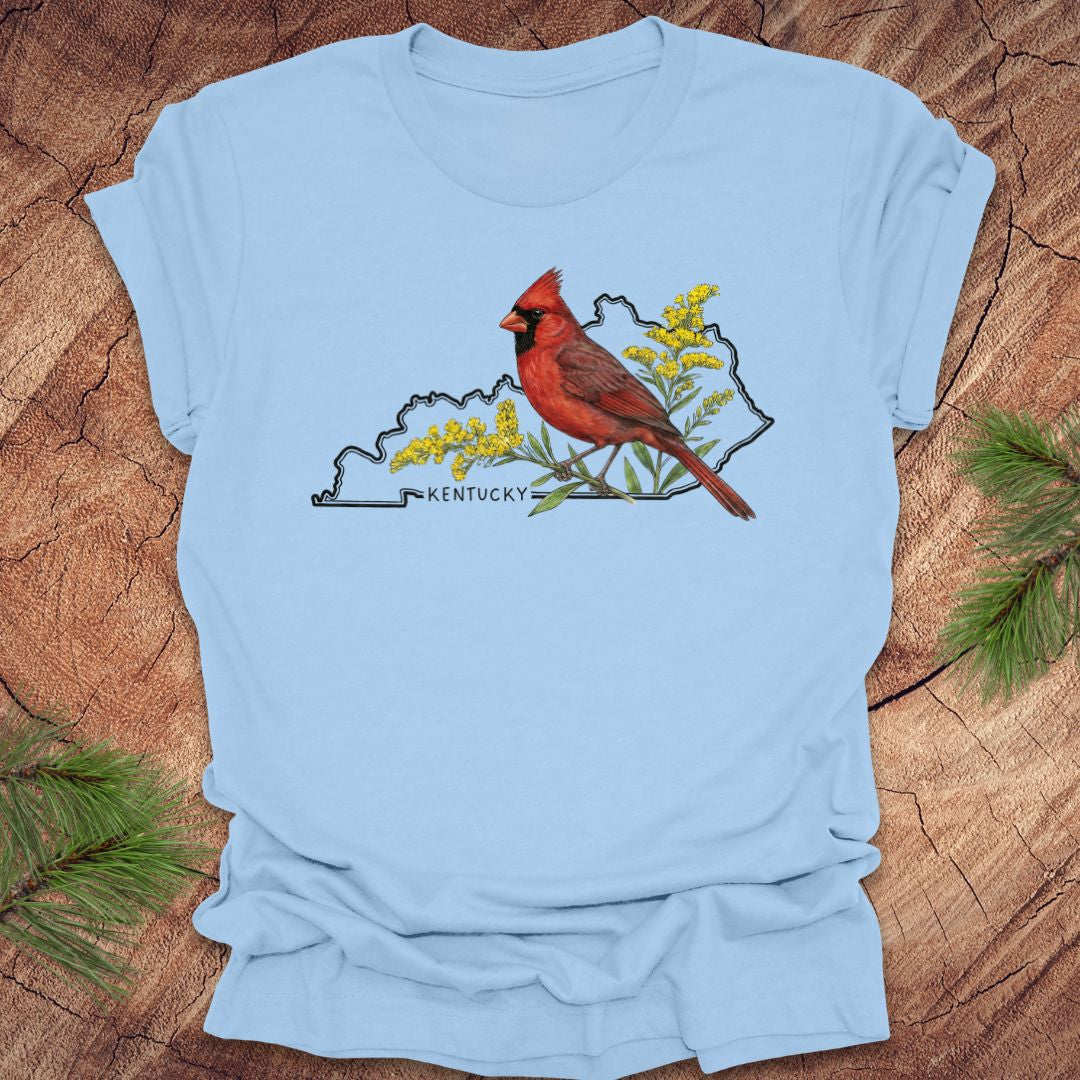 Baby blue t-shirt with the outline of the state of kentucky and state bird cardinal and goldenrod design on a wooden surface