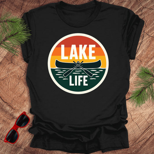 A black t-shirt with a vintage sunset design featuring a canoe and the text 'LAKE LIFE'. The shirt is presented on a wooden surface with pine needles and a pair of sunglasses to the side.