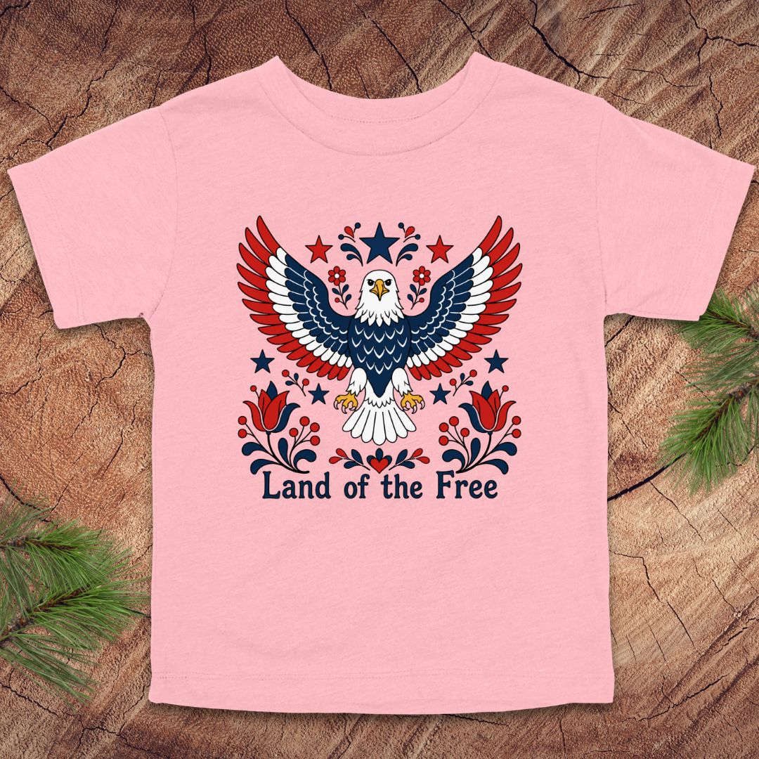 Pink t-shirt with an eagle design and 'Land of the Free' text on a wooden surface.