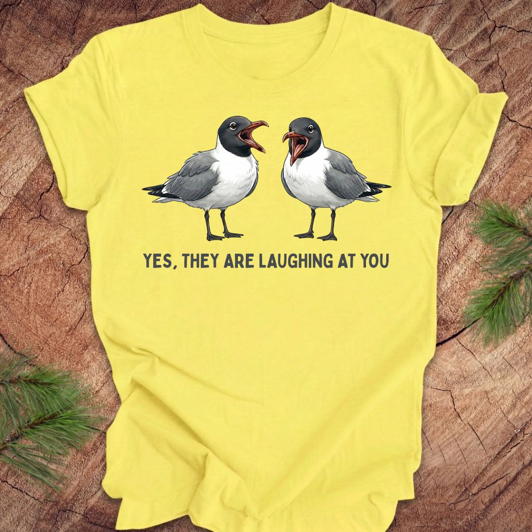 Yellow t-shirt with two laughing gulls and text, "yes, they are laughing at you" on a wood background.
