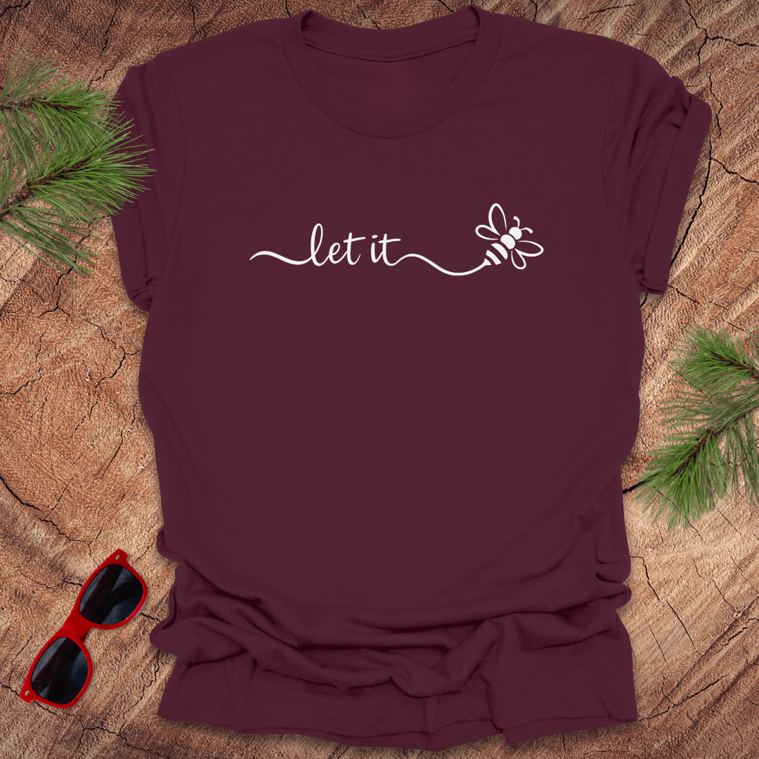 Maroon t-shirt with 'let it' and a bee design on a wooden surface with sunglasses and greenery.