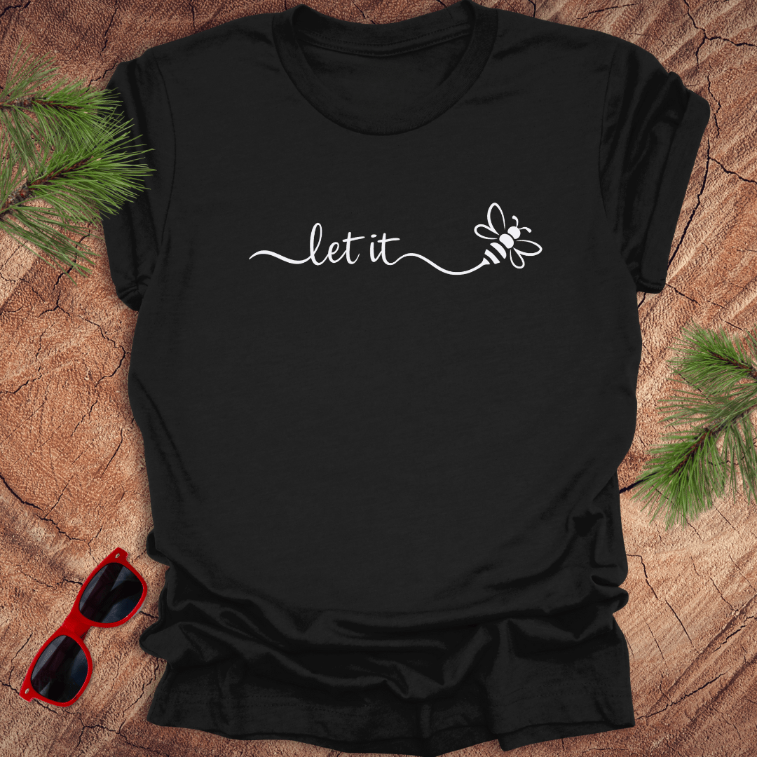 Black t-shirt with 'let it' and a bee design on a wooden surface with sunglasses and pine branches.