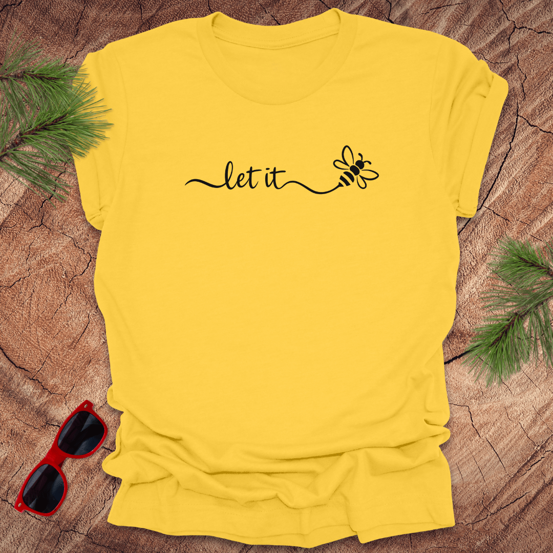 Yellow t-shirt with 'let it' and bee design on a wooden surface with sunglasses and greenery.