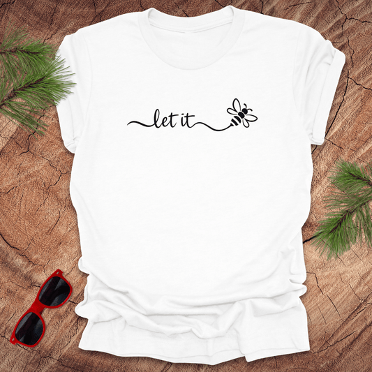 White t-shirt with 'let it' text and bee graphic on a wooden surface with sunglasses and greenery.