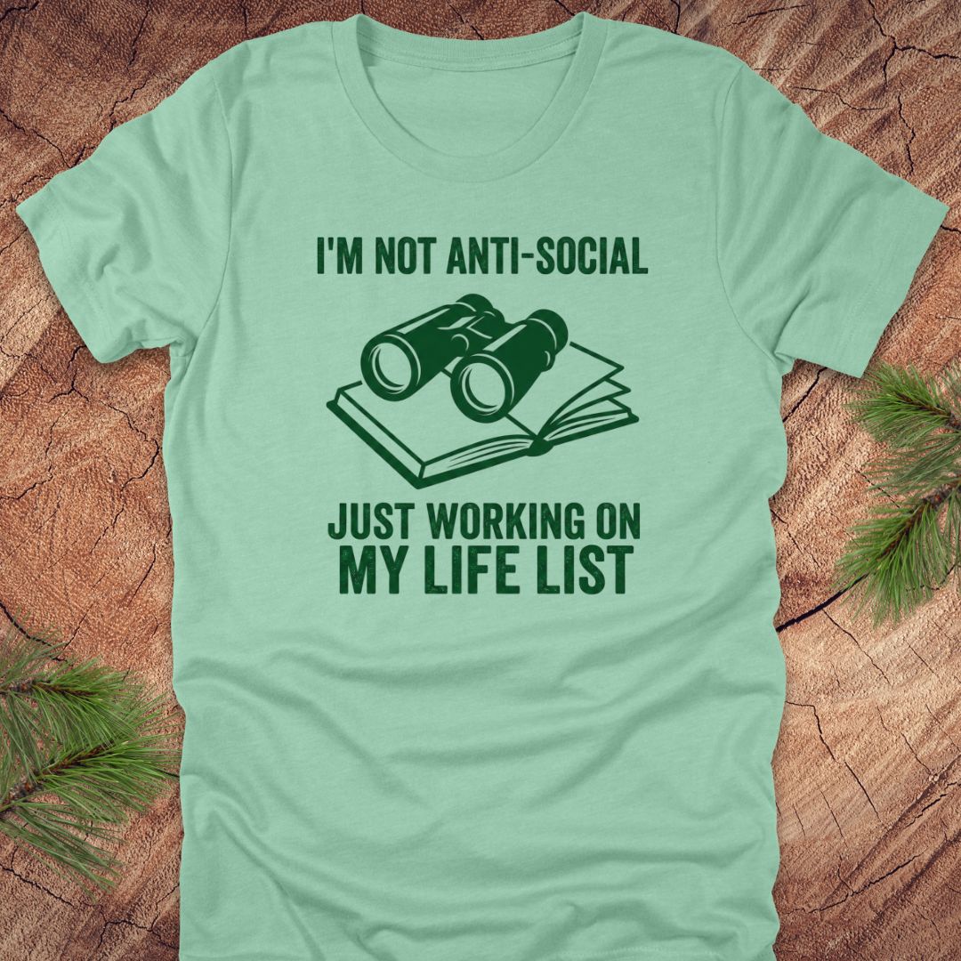 Mint green t-shirt with green text 'i'm not anti-social just working on my life list' and binoculars graphic on a wooden surface