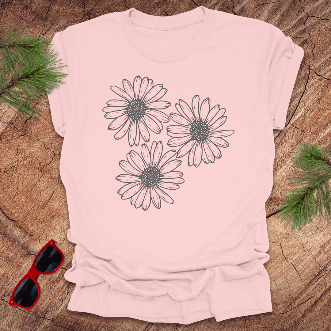 A pink t-shirt with 3 daisies prominently displayed. on a wooden surface with sunglasses and greenery.