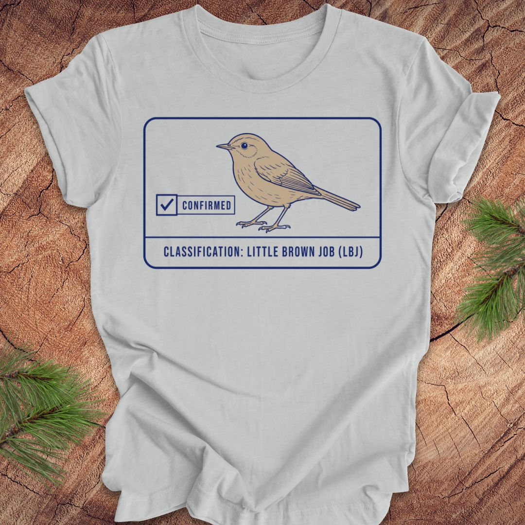 A gray short-sleeve crewneck t-shirt laid flat on a rustic wood background with pine needles in the corners. The shirt features a graphic of a little brown job bird with a 'Confirmed' checkmark and the text 'CLASSIFICATION: LITTLE BROWN JOB (LBJ)'.