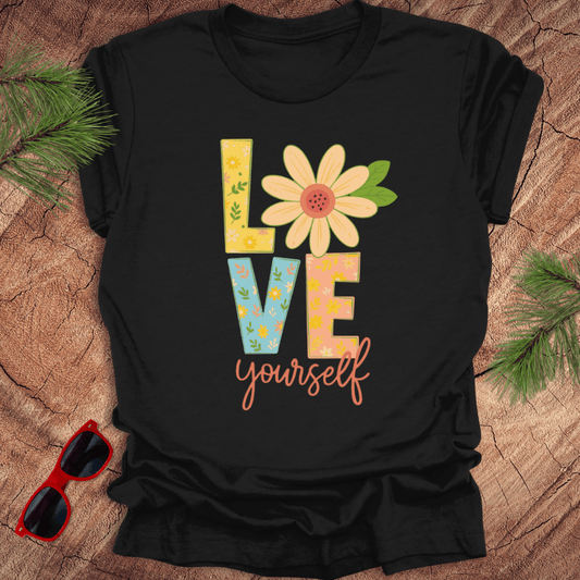 Black t-shirt with 'Love yourself' design on a wooden surface with sunglasses and greenery.