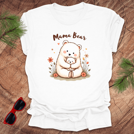 A white t-shirt with a graphic of a mama bear holding a cub, accompanied by boho floral details and the text 'Mama Bear'. The shirt is presented on a wood surface with pine needles sunglasses.