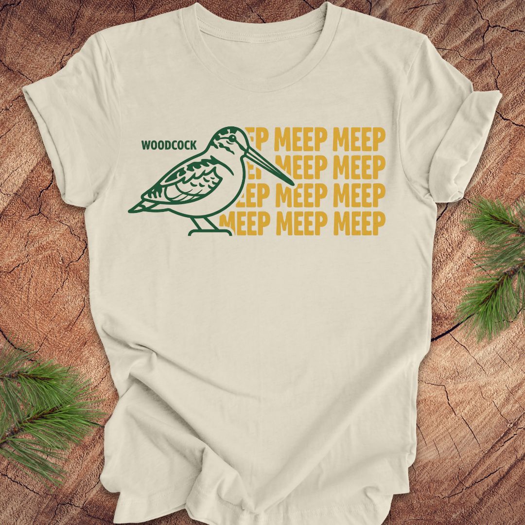 Beige t-shirt with green woodcock graphic and 'MEEP MEEP MEEP' text on a wooden surface.
