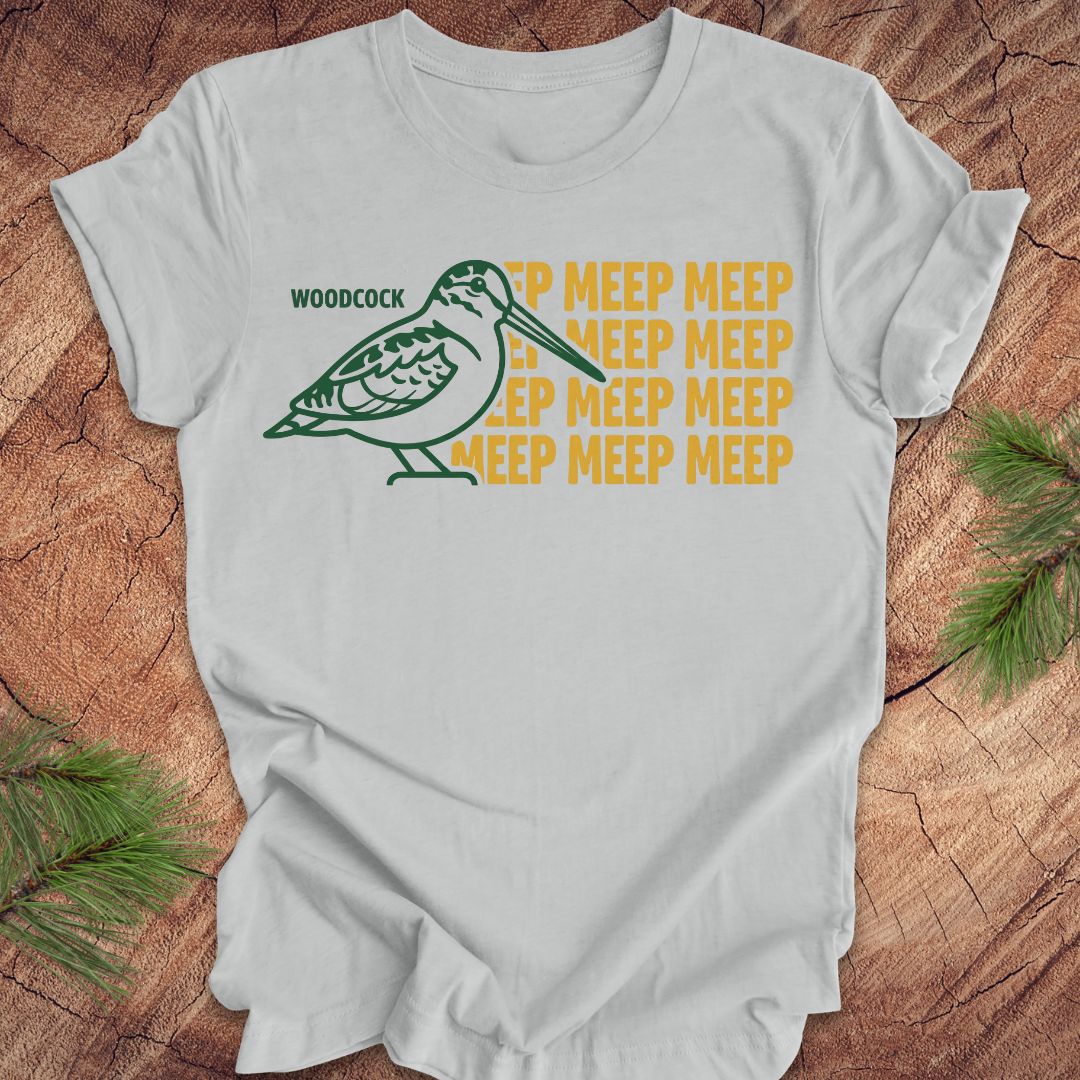 Gray t-shirt with a green woodcock graphic and yellow 'meep' text on a wooden surface.