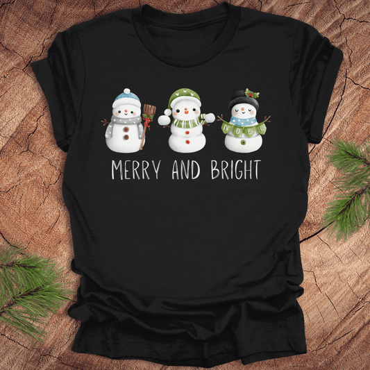 Black t-shirt with snowman design and 'Merry and Bright' text on a wooden surface.