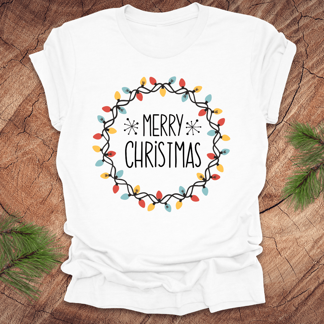 White t-shirt with 'Merry Christmas' design on a wooden surface with pine branches.