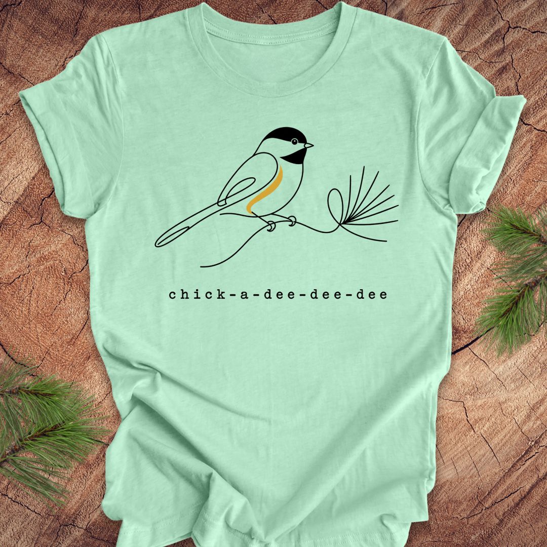 Light green t-shirt with a chickadee design and 'chick-a-dee-dee-dee' text on a wooden surface.