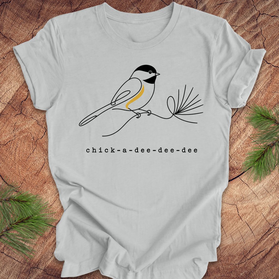 Gray t-shirt with chickadee design and 'chick-a-dee-dee-dee' text on a wooden surface.