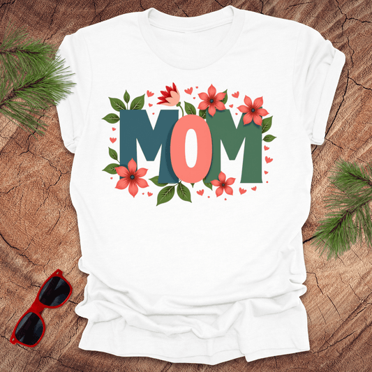 white tshirt with the text 'mom' in bold colorful letters surrounded by flowers, on a wooden background with pine needles and a pair of sunglasses.