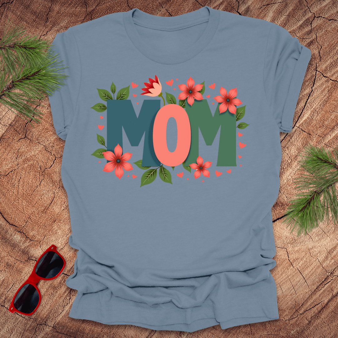 steel blue tshirt with the text 'mom' in bold colorful letters surrounded by flowers, on a wooden background with pine needles and a pair of sunglasses.