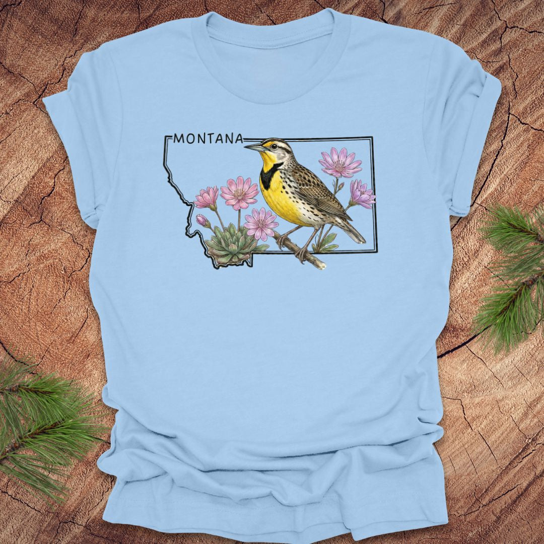 Baby blue t-shirt with the state outline of Montana with a graphic of a western meadowlark and bitteroot, on a wooden surface