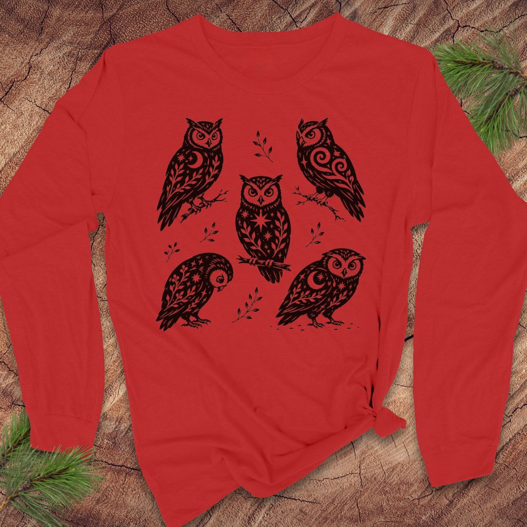 Red long-sleeve t-shirt with black folk-art owls decorated with crescent moons, stars, and vines, laid flat on a rustic wood background with pine branches.