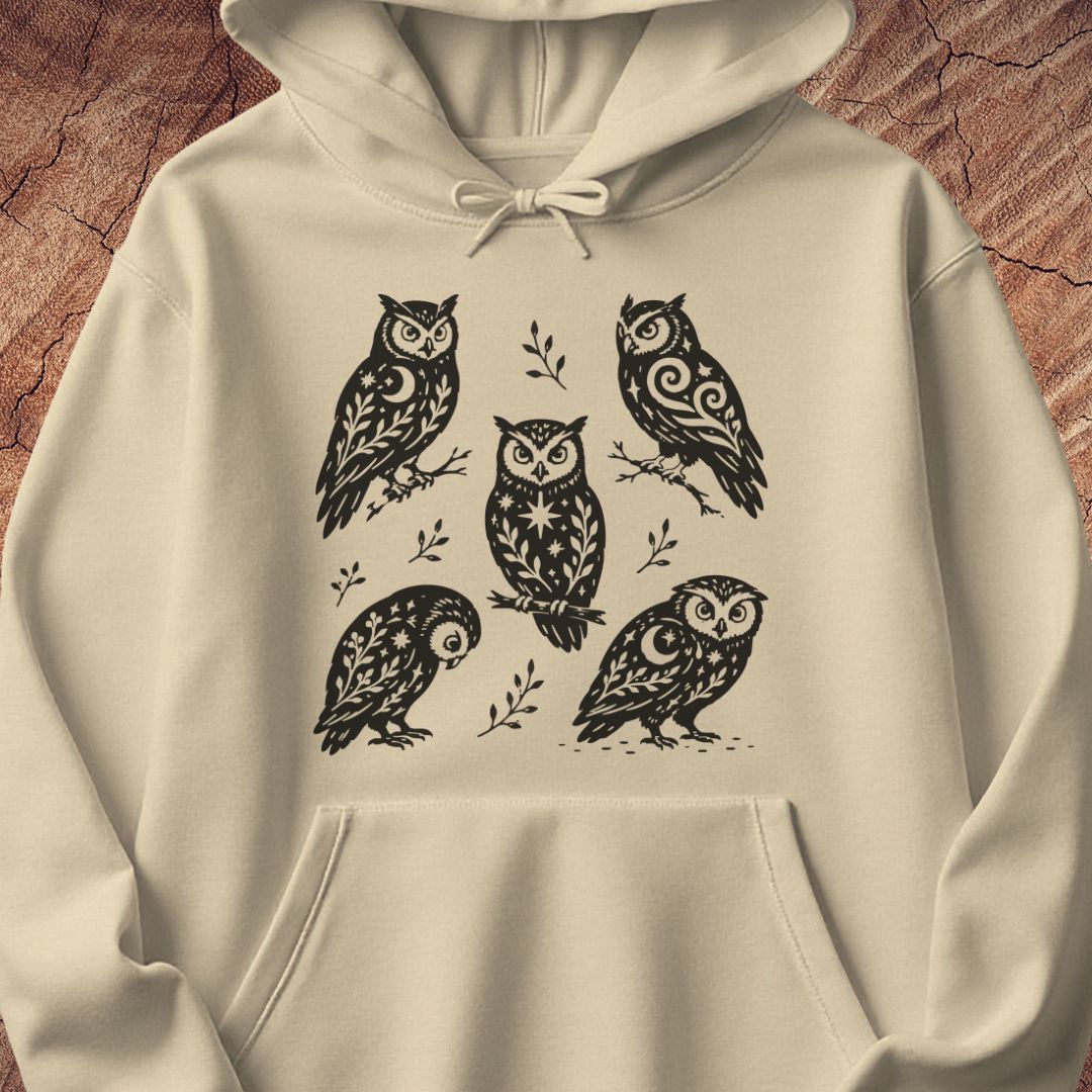 Sand beige hoodie with black folk-art owls decorated with crescent moons, stars, and vines, laid flat on a rustic wood background.