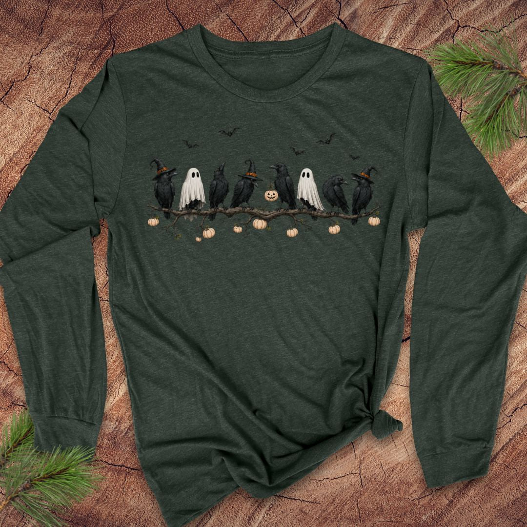 A dark forest green long-sleeve t-shirt with a Halloween graphic print of crows, ghosts, pumpkins, and bats, laid flat on a rustic wood background with pine branches.