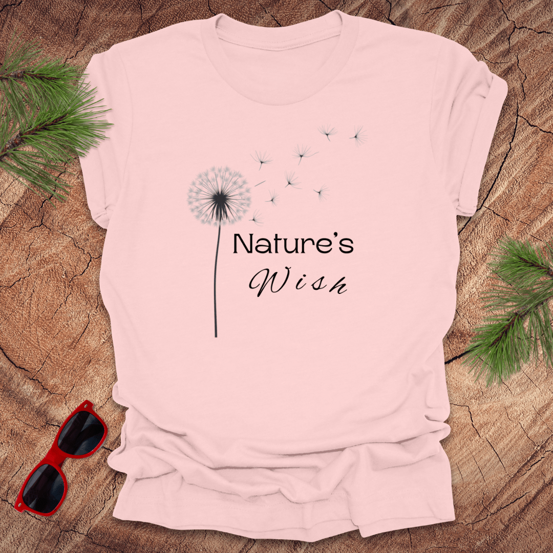 pink t-shirt with a dandelion seed design and the text 'Nature's Wish' on the front, presented on a wooden surface with pine needles and red sunglasses.